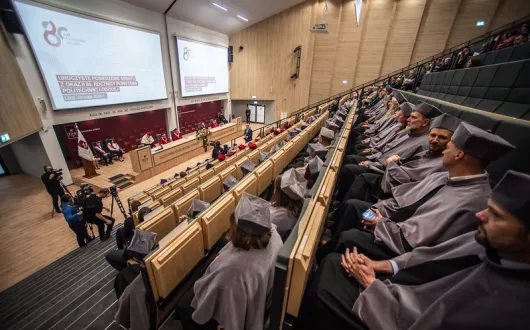 członkowie senatu ubrani w tradycyjne togi koloru szarego, przed nimi telebim z wyświetlonym slajdem z okazji 80-lecia politechniki Łódzkiej