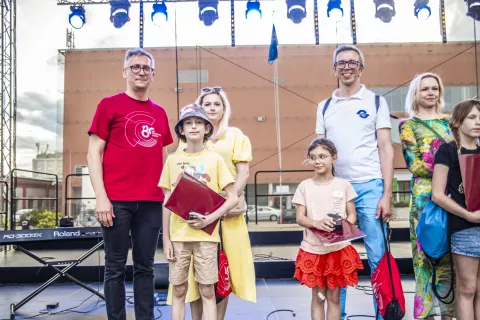 dzieci i opiekunowie w kolorowych ubraniach stoją na scenie, za nimi reflektory